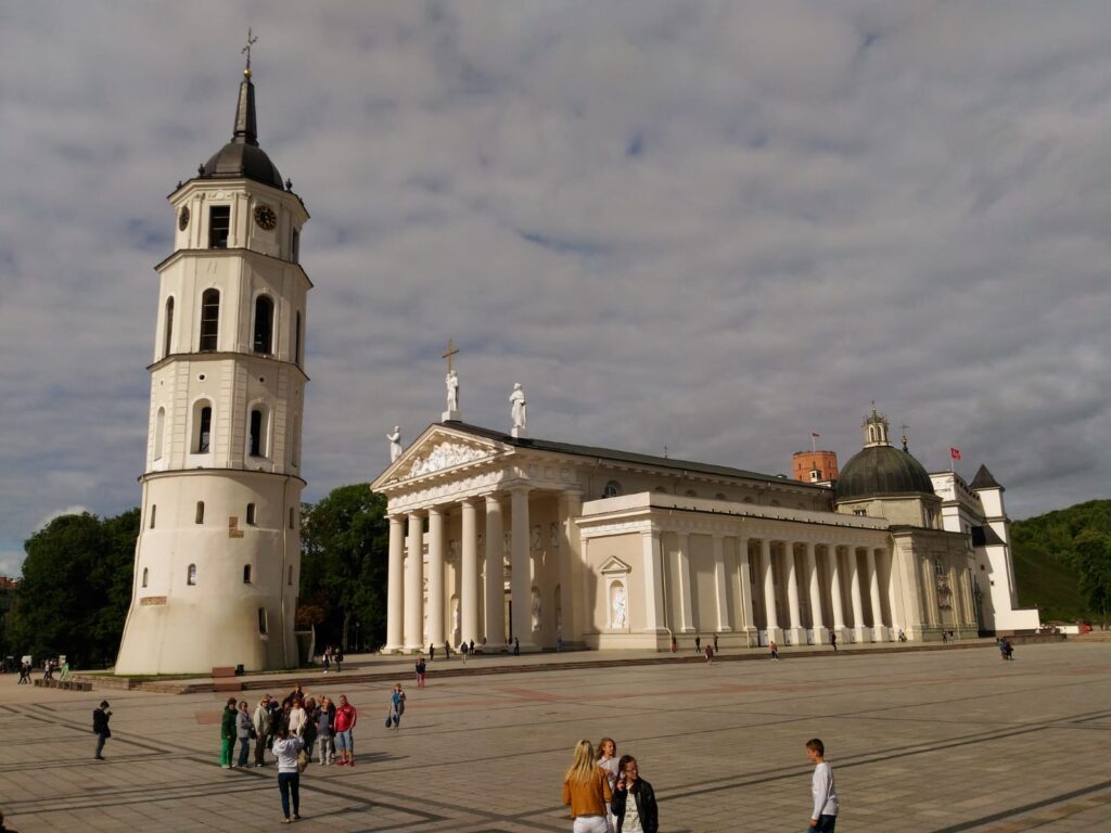Plac z dużym, białym kościołem. Kościół z przodu i boku ma liczne kolumny. Przed wejściem wysoka biała wieża dzwonnica.