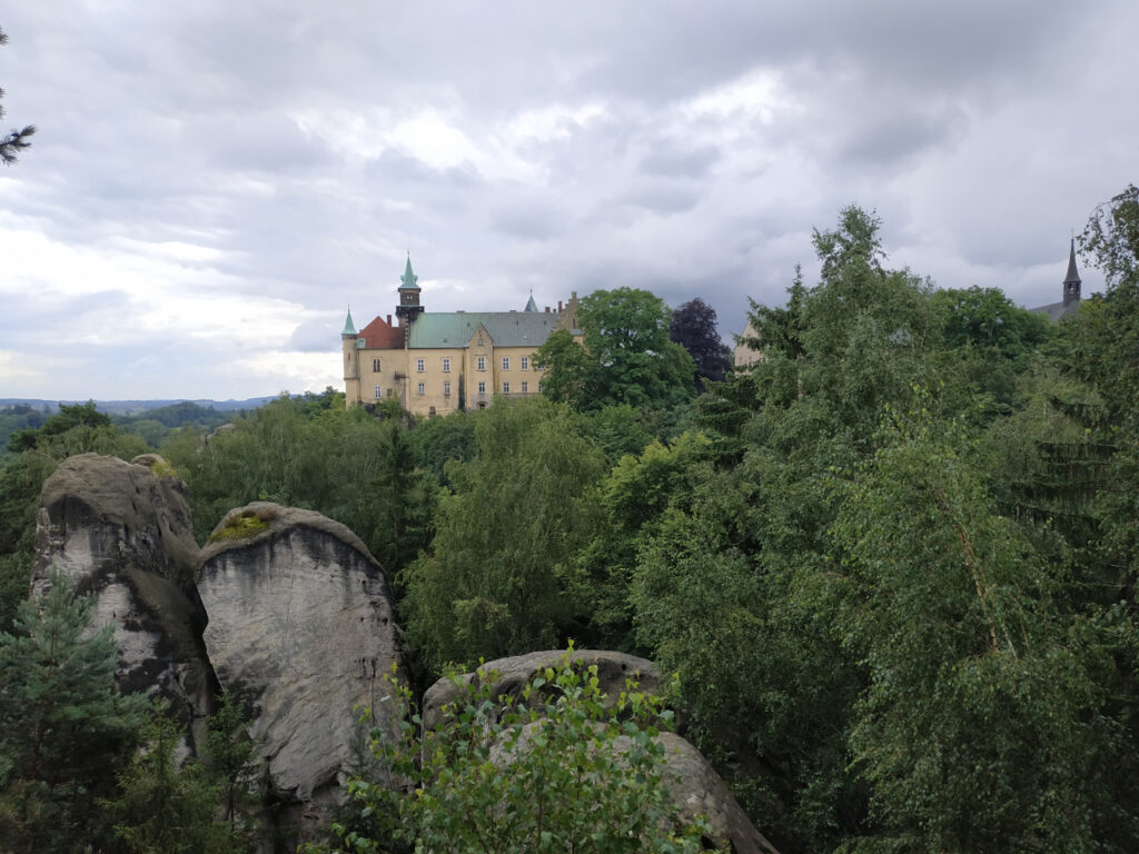 Widok na czeski zamek Gruba Skała. Na pierwszym planie trochę drzew i kilka skał.