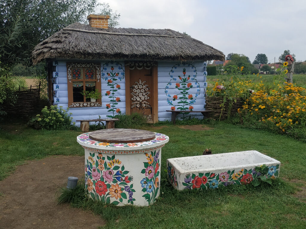 Zalipie, malowana wieś Nieduży budynek kryty strzechą a bliżej studnia i ławka. Wszystko pomalowane w kwiaty